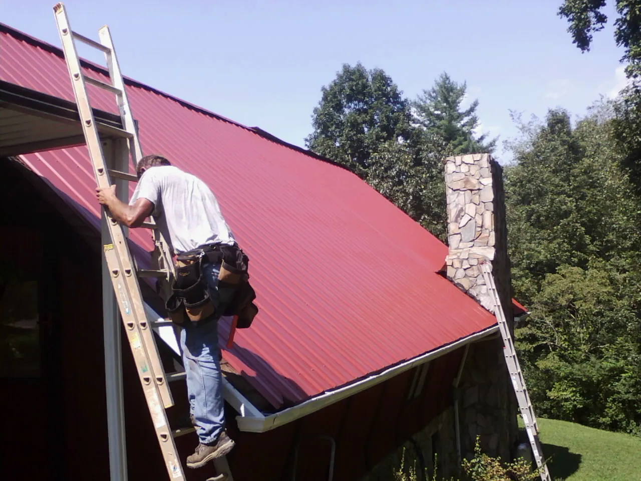 Roofer on a ladder beside a red metal roof and stone chimney — Soffit & Fascia Repair by Roofing Brothers in Cliffside Park